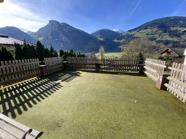 A fenced yard with a green lawn, surrounded by trees. Majestic mountains and blue sky in the background, sunlight casting strong shadows over the ground. Peaceful setting.
