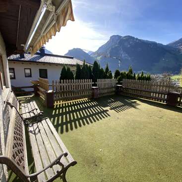 Eine sonnige Terrasse mit Holzbänken und einem Lattenzaun blickt auf eine malerische Berglandschaft. Der Schattenwurf auf dem Kunstrasen schafft eine friedliche, einladende Atmosphäre.