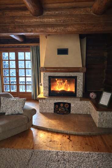 Le salon chaleureux de cette cabane en rondins est doté d'une cheminée crépitante, d'un canapé confortable orné de coussins à motifs floraux, de poutres apparentes rustiques, de grandes fenêtres et d'une vue sur un paysage enneigé. Une atmosphère chaleureuse.