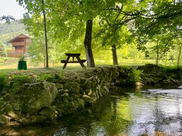 Une scène paisible au bord d'une rivière, avec une table de pique-nique en bois sous des arbres verdoyants, près d'un chalet rustique. Un talus en pierre, des eaux calmes, une atmosphère champêtre sereine.