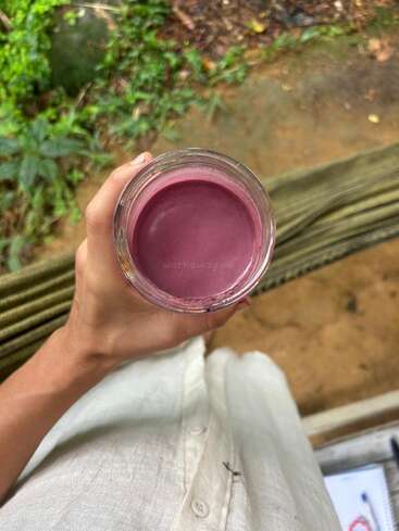 A person holds a glass of purple smoothie or juice, standing outdoors on a green hammock, wearing a light-colored shirt. The background is earthy and lush.