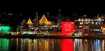 Una ciudad a orillas del río por la noche, con los edificios iluminados con luces verdes, amarillas y rojas. Un árbol de Navidad decorado destaca en primer plano, con reflejos de colores que brillan sobre el agua.
