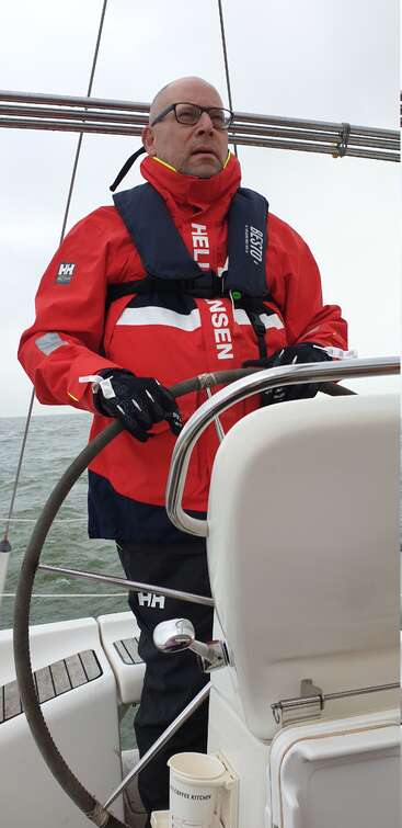 Un hombre con una chaqueta Helly Hansen y un chaleco salvavidas se encuentra al timón de un velero, navegando por aguas grises y agitadas bajo un cielo nublado.
