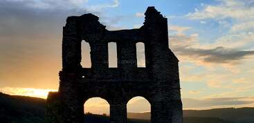 Las antiguas ruinas de piedra se recortan contra un cielo crepuscular lleno de colorido, con nubes teñidas de naranja y azul. Las colinas onduladas y los árboles en la lejanía completan esta tranquila estampa.