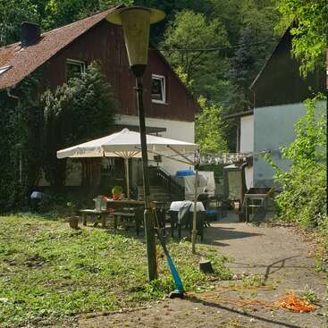 Uma cena aconchegante no quintal, com uma casa de madeira, um guarda-sol, uma mesa de piquenique, ferramentas de jardinagem e varal. Cercada por uma vegetação exuberante e luz natural.