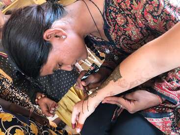 The image depicts a woman with dark hair and a black top, leaning over to apply henna to another person\'s hand, surrounded by other people.