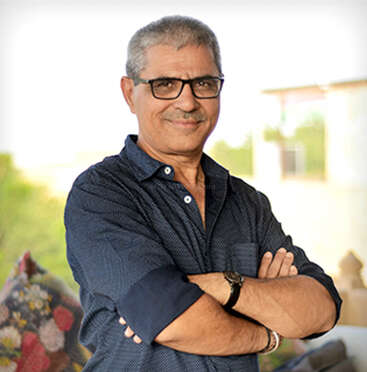 L'image représente un homme d'âge moyen aux cheveux gris courts, vêtu d'une chemise bleu foncé et debout, les bras croisés, sur un fond extérieur flou.