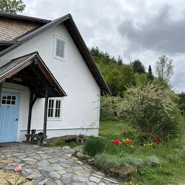 L'image représente une charmante maison blanche avec une porte bleue, nichée au milieu d'une verdure luxuriante et de fleurs éclatantes, sur fond de ciel nuageux.