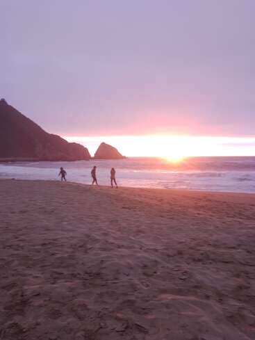 Three people walk along a sandy beach at sunset, with waves gently crashing and distant rocky hills silhouetted against a vibrant, glowing horizon and purple sky.