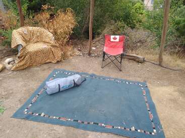 A cozy outdoor setup with a leopard-print covered chair, Canadian flag folding chair, large green rug, and camping gear, surrounded by greenery and dry soil.