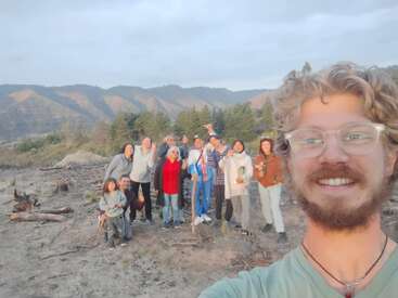 Un grupo de personas sonrientes posa al aire libre en un paisaje natural y montañoso. Un hombre con gafas se hace un selfi, capturando a todos mientras disfrutan del hermoso y soleado paisaje.