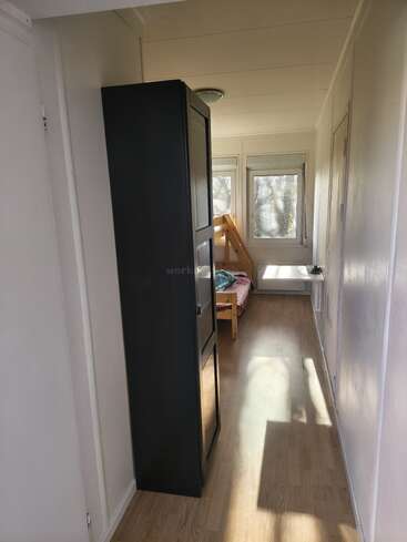 Una habitación estrecha y luminosa, con suelo de madera, un armario alto y negro, una litera junto a unos grandes ventanales y paredes blancas que crean un ambiente luminoso y acogedor.