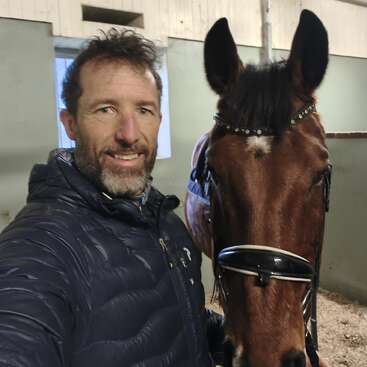 Un hombre sonriente, vestido con una chaqueta azul marino, se hace un selfi con un caballo castaño en un establo. El caballo lleva una brida y una testera decorada.