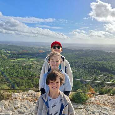 Three children stand in a row on a rocky hilltop, smiling. Behind them, lush green forests and fields stretch under a bright blue, partly cloudy sky.