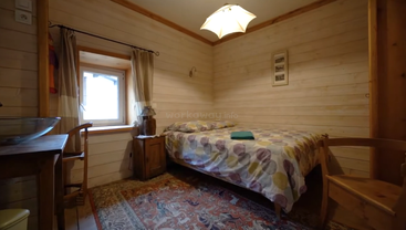 This cozy wooden room features a comfortable bed with colorful bedding, a window with light curtains, a small desk, wooden furniture, and a patterned rug.