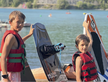 Two boys in red life vests stand by a lake holding surfboards. The weather is sunny. Water and trees are visible in the background. They look ready for adventure.