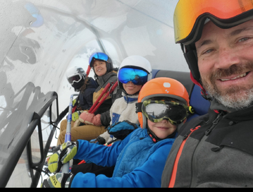 A happy group of five people, dressed in colorful ski gear, sit closely together on a ski lift under a protective cover, smiling and ready for adventure.