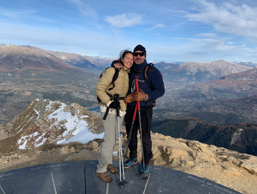 A smiling couple in hiking gear stands on a mountain summit, trekking poles in hand. Snow-capped peaks and a breathtaking valley stretch out behind them.