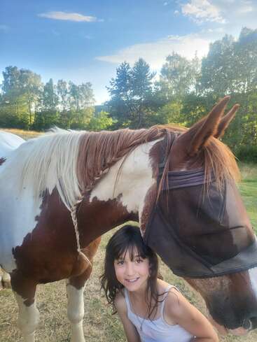 A young girl smiles while standing beside a brown and white horse wearing a fly mask, surrounded by lush trees under a bright, partly cloudy sky.