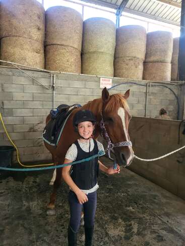 A smiling child in riding gear stands next to a saddled brown horse inside a stable. Large hay bales are stacked behind them against the wall.
