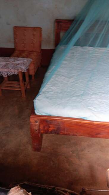 Esta imagen muestra un dormitorio sencillo con una cama de madera cubierta por una mosquitera azul, una silla naranja, una mesita y un fondo de pared liso.