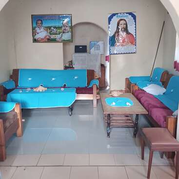 La imagen muestra un salón ordenado con sofás de madera cubiertos de tela azul, una mesa de centro, carteles religiosos en la pared, suelo de baldosas y un ambiente tranquilo y acogedor.