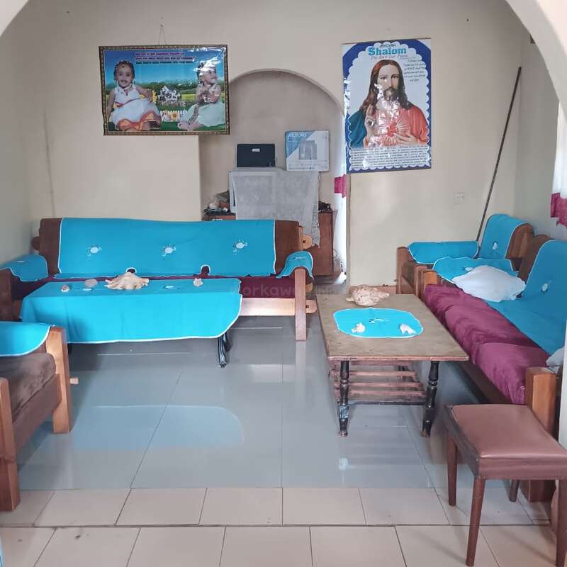 La imagen muestra un salón ordenado con sofás de madera cubiertos de tela azul, una mesa de centro, carteles religiosos en la pared, suelo de baldosas y un ambiente tranquilo y acogedor.