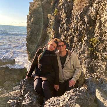 The image depicts a couple sitting on rocks by the ocean, with the man wearing a beige jacket and the woman in a black sweater, set against a rocky cliff backdrop.