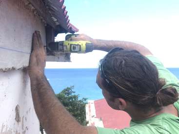Un homme aux cheveux bruns coiffés en chignon utilise une perceuse électrique jaune sur le côté d'un bâtiment, avec un plan d'eau et des arbres en arrière-plan.