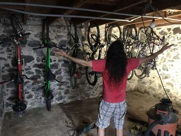 Un homme aux cheveux longs se tient dans un garage, les bras tendus, entouré de vélos et d'outils, vêtu d'une chemise rouge et d'un short à carreaux.