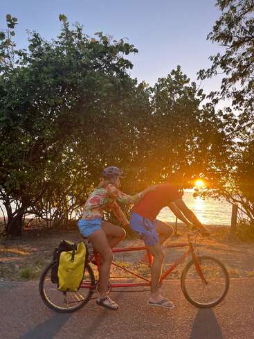 A couple rides a tandem bicycle along a sunlit road by the beach. Surrounded by trees, the golden sunset creates a warm, peaceful atmosphere.