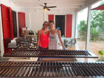 Two smiling women stand together on a porch with red shutters, working on refurbishing wooden window blinds. The atmosphere is bright, casual, and industrious.