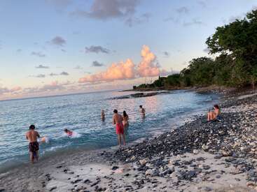 A group of people enjoy swimming and relaxing on a rocky beach at sunset, with beautiful clouds, calm blue water, and trees along the shore.