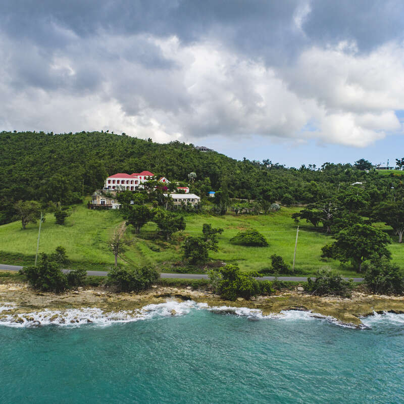 L'image représente un paysage côtier serein, avec une grande maison blanche au toit rouge, nichée dans une végétation luxuriante et surplombant l'océan.