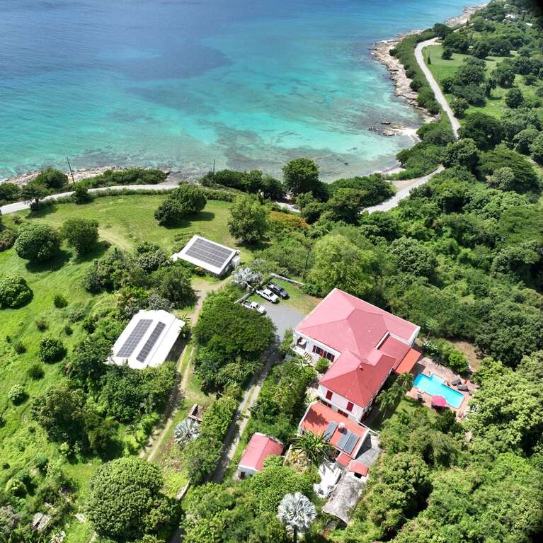 This aerial image shows a coastal estate with red-roofed buildings, solar panels, lush greenery, a swimming pool, clear turquoise water, and a winding seaside road.