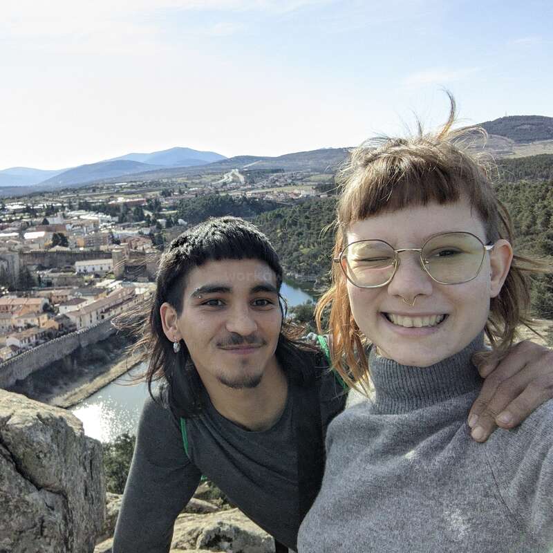 Un couple souriant prend un selfie au sommet d'un promontoire rocheux, avec en arrière-plan, sous un ciel dégagé, une ville pittoresque, une rivière sinueuse, des collines boisées et des montagnes au loin.