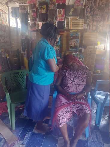 In a small salon, a woman styles another woman's hair. Hair extensions hang on the walls, while the seated client wears a pink patterned dress.
