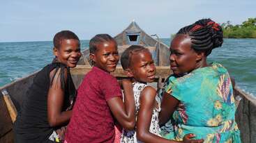 A imagem mostra três mulheres e uma criança em frente a um barco de madeira na água, em um céu azul sereno.