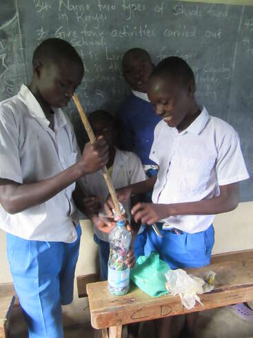 The image shows four children in a classroom, standing around a table with a plastic bottle filled with small items and a stick inserted into it.
