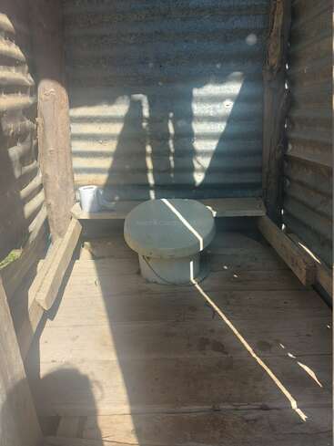 This image shows a simple outdoor toilet with a wooden floor, corrugated metal walls, a basic toilet seat, a roll of toilet paper, and sunlight streaming in.