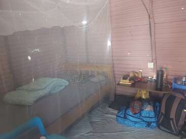 This image shows a simple room with a bed covered by a mosquito net, a desk with essentials, a backpack, bags, and a soft pink corrugated wall.