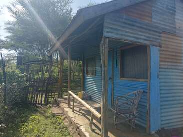 This image shows a simple, rural house made from corrugated metal sheets, with a small porch, wooden posts, outdoor chair, and surrounded by greenery and trees.