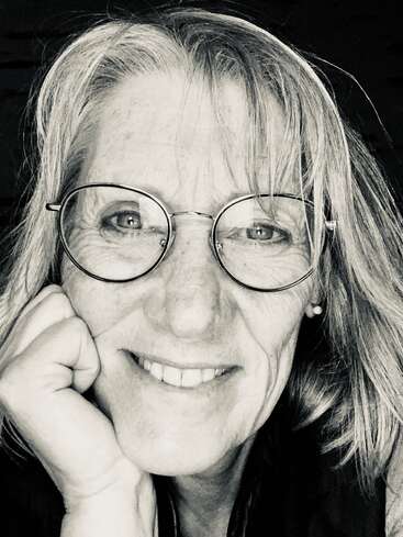 Das Bild ist eine Schwarz-Weiß-Nahaufnahme einer Frau mit langen Haaren, die eine Brille trägt und in die Kamera lächelt, mit der Hand im Gesicht.