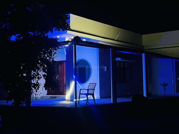Le porche d'une maison moderne, éclairé de nuit par des lumières bleues, présente une chaise unique, une fenêtre circulaire, une silhouette de feuillage et crée une atmosphère paisible et accueillante.