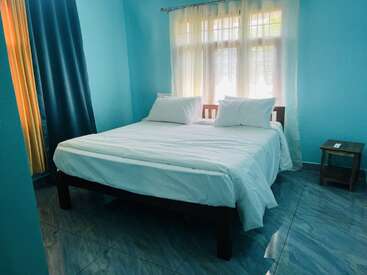 L'image montre une chambre propre et simple aux murs bleus, un grand lit avec des draps blancs, deux oreillers, la lumière du soleil à travers les rideaux et une table de nuit en bois.