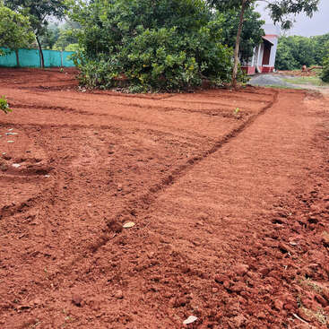 Freshly tilled red soil field with visible furrows, surrounded by green trees and a distant small building. Area looks prepared for planting or gardening.