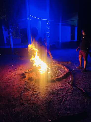 A vibrant outdoor campfire burns brightly at night, surrounded by stones. Blue and purple lighting illuminate the scene as people stand around, enjoying the warmth.