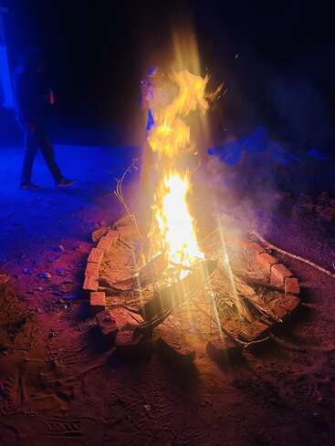 A bright bonfire burns intensely, surrounded by bricks and dry wood. Two people are nearby, illuminated by blue lighting, creating a lively nighttime outdoor atmosphere.