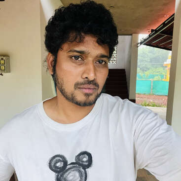 A man with curly hair and a beard stands indoors wearing a white t-shirt with a bear face design, posing for a selfie with a neutral expression.