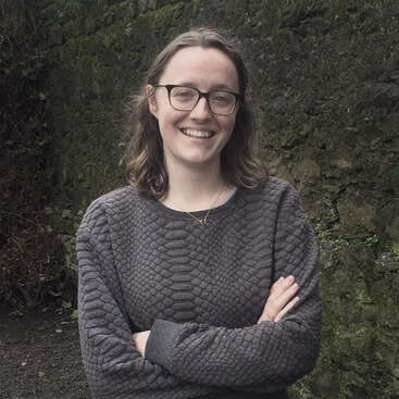 A woman with glasses, wearing a textured gray sweater and a necklace, stands outdoors smiling, arms crossed, with a mossy stone wall in the background.